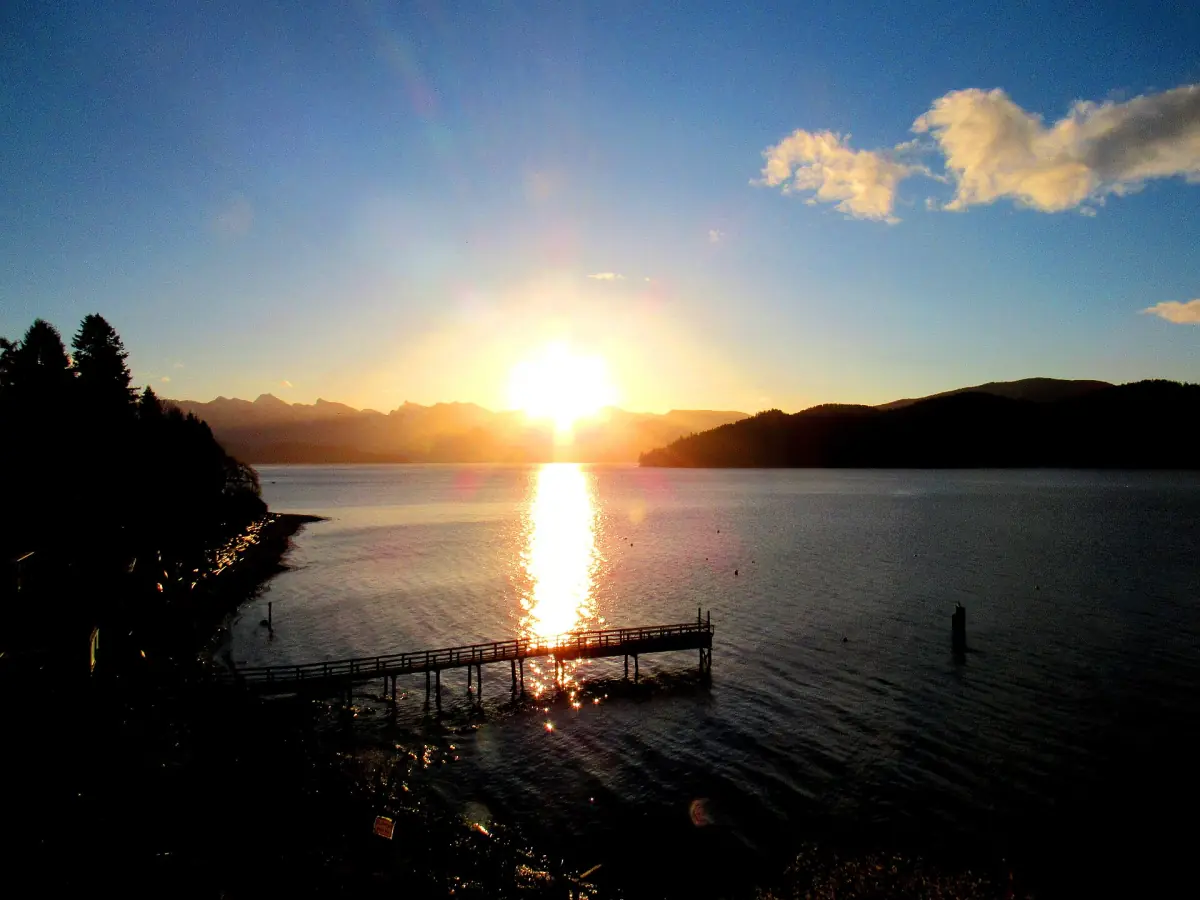 Sun setting behind mountains over a calm inlet with a small wooden dock